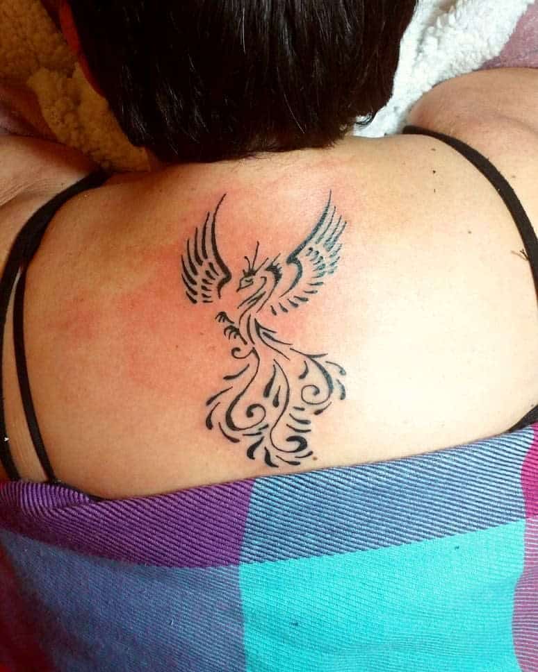 A black phoenix rising tattoo on a person's upper back