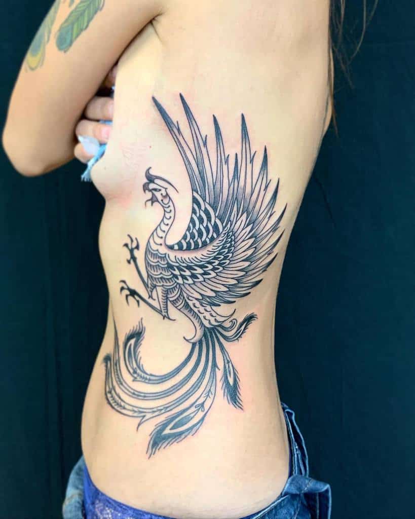 Tattoo of a phoenix rising on the side of a person's torso, wings spread and tail feathers flowing
