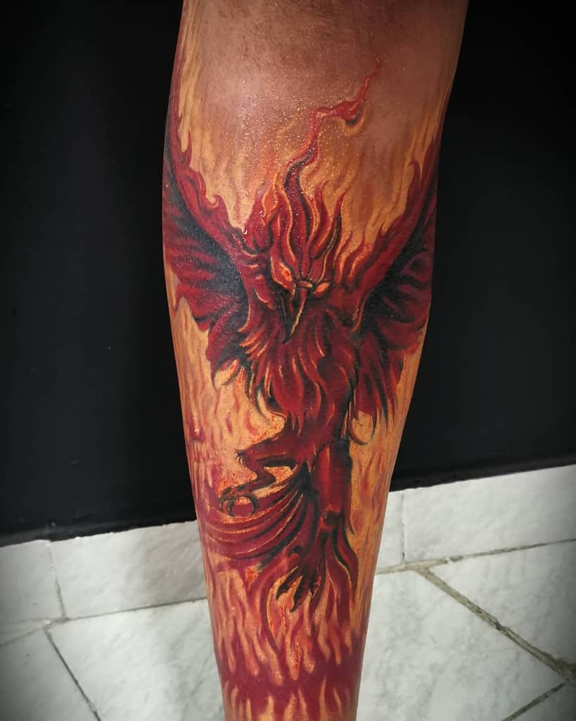 A tattoo of a fiery red phoenix with wings spread, rising from flames on a calf