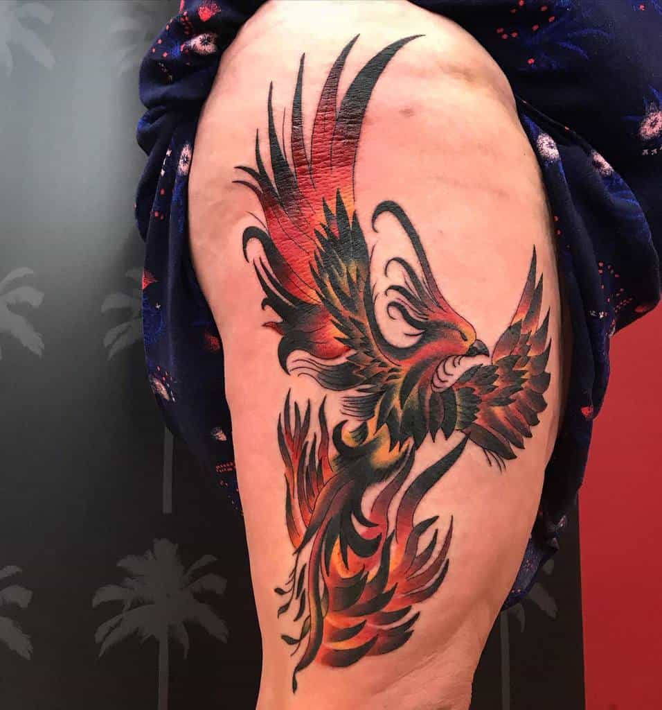 Tattoo of a fiery phoenix rising in vibrant red, orange, and black ink on an upper thigh