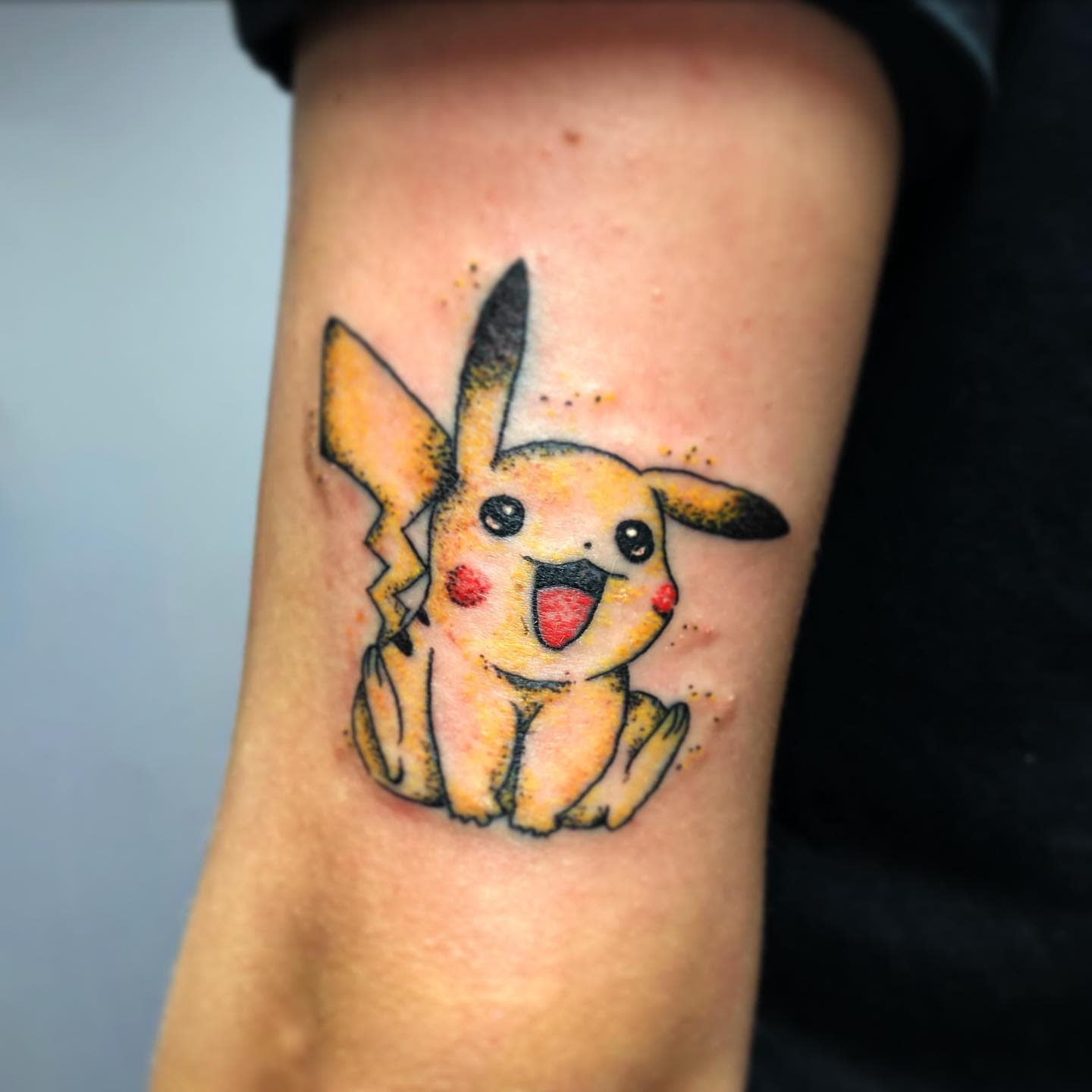 Tattoo of Pikachu on an arm, smiling and sitting with bright colors and shading