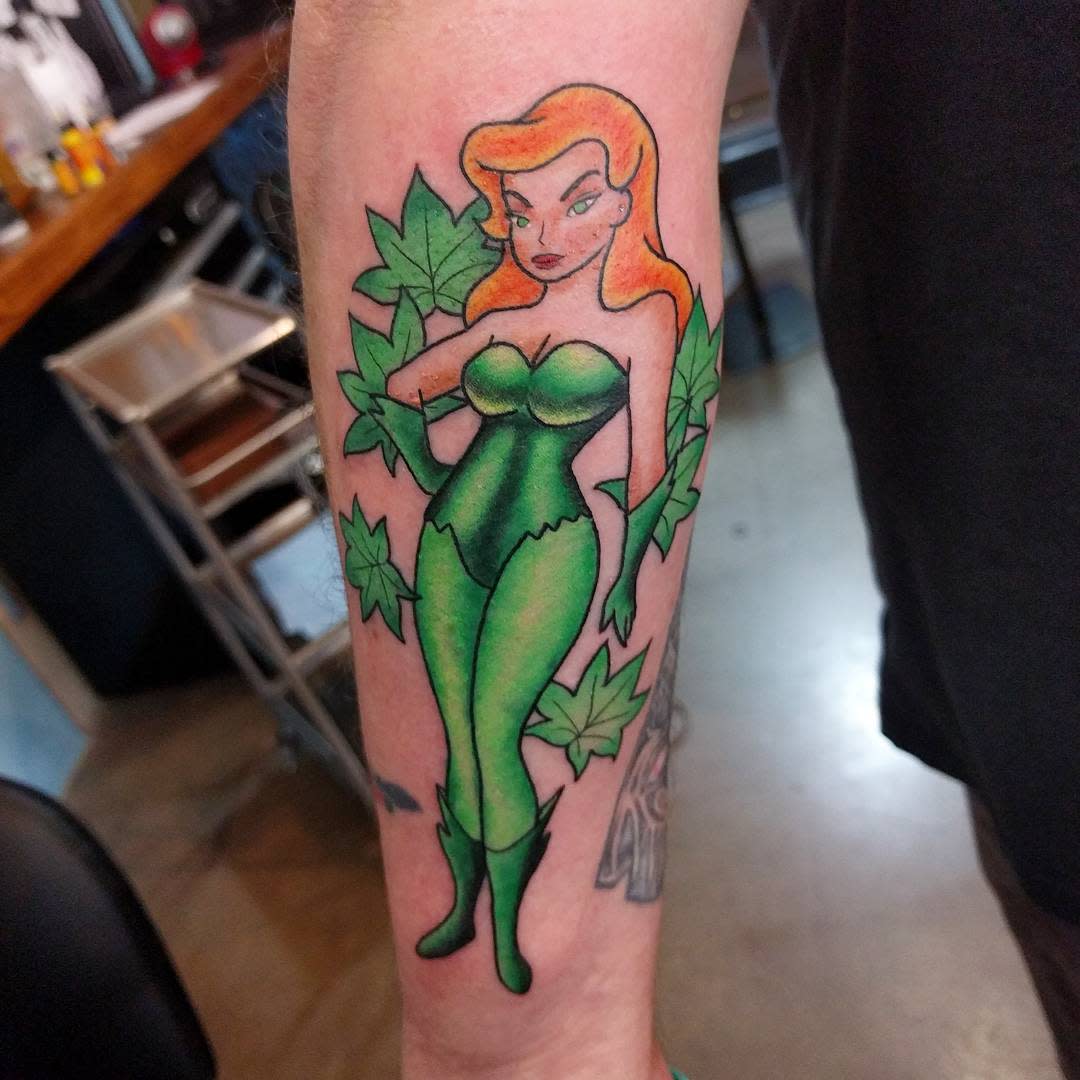 Tattoo of a woman in a green outfit surrounded by poison ivy leaves on a forearm