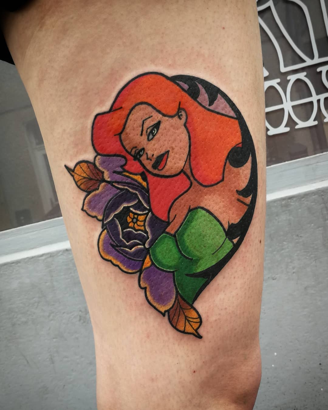 Tattoo of a woman with red hair, green top, and purple flowers on the thigh
