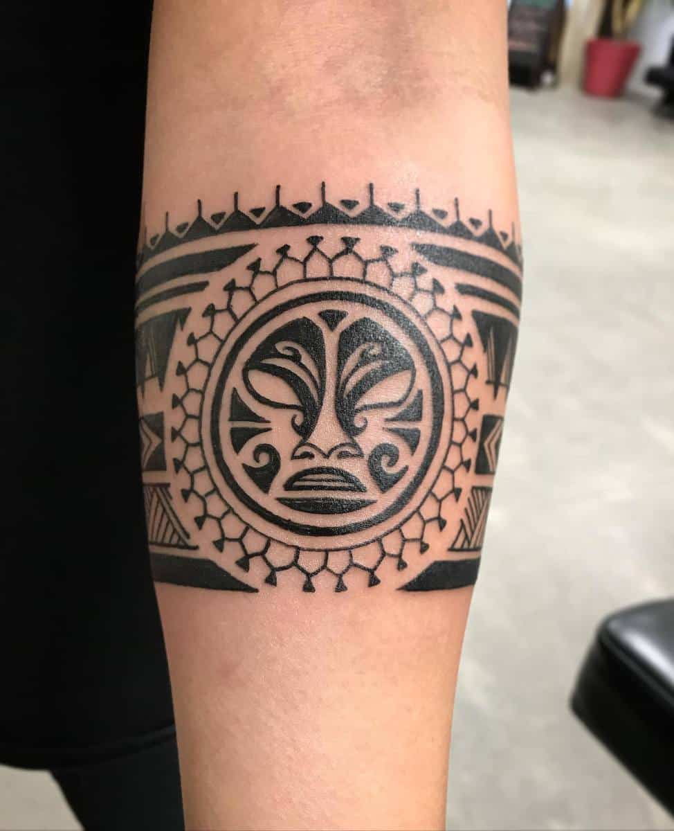 Forearm tattoo with a circular Polynesian tribal design, central figure, and intricate geometric patterns