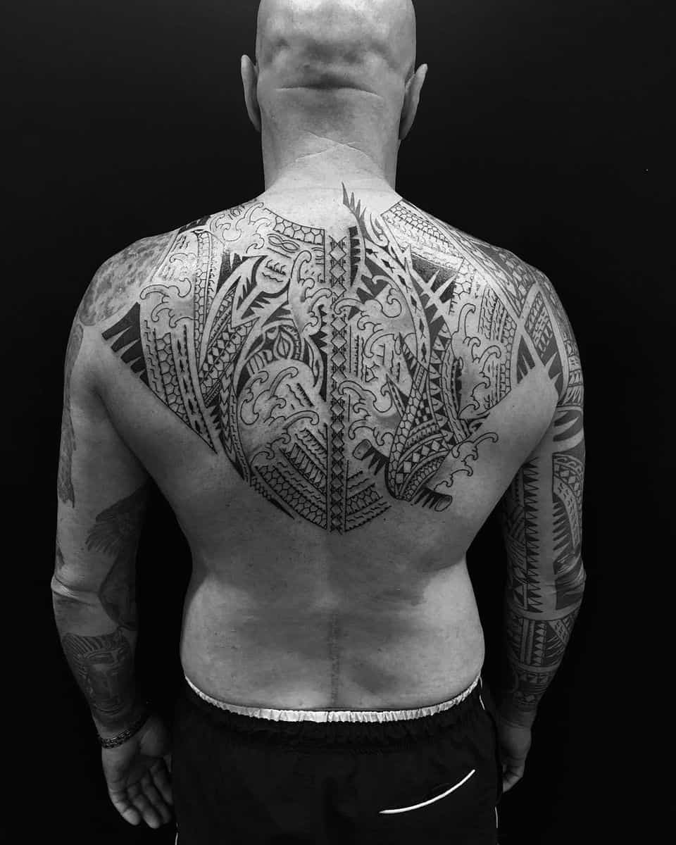 A bald man with intricate Polynesian tribal tattoos covering his back and arms against a black background