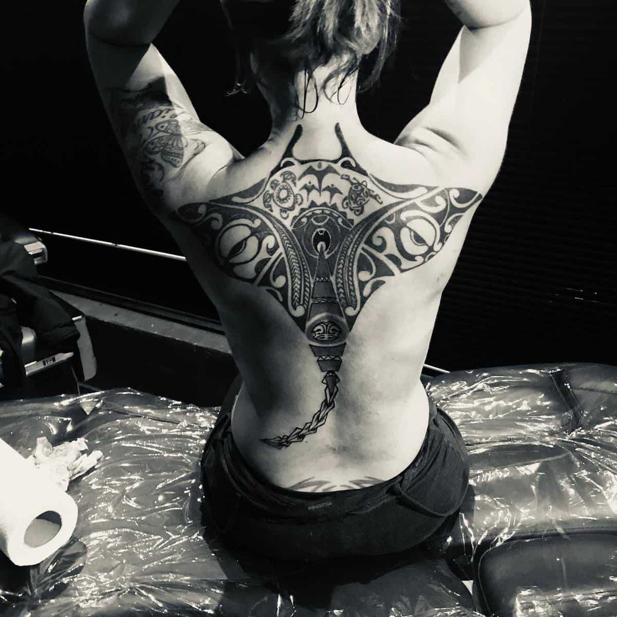 A figure with an intricate back tattoo showing a stylized manta ray, sitting with arms raised, featuring captivating Polynesian patterns that add cultural depth and elegance to the artwork