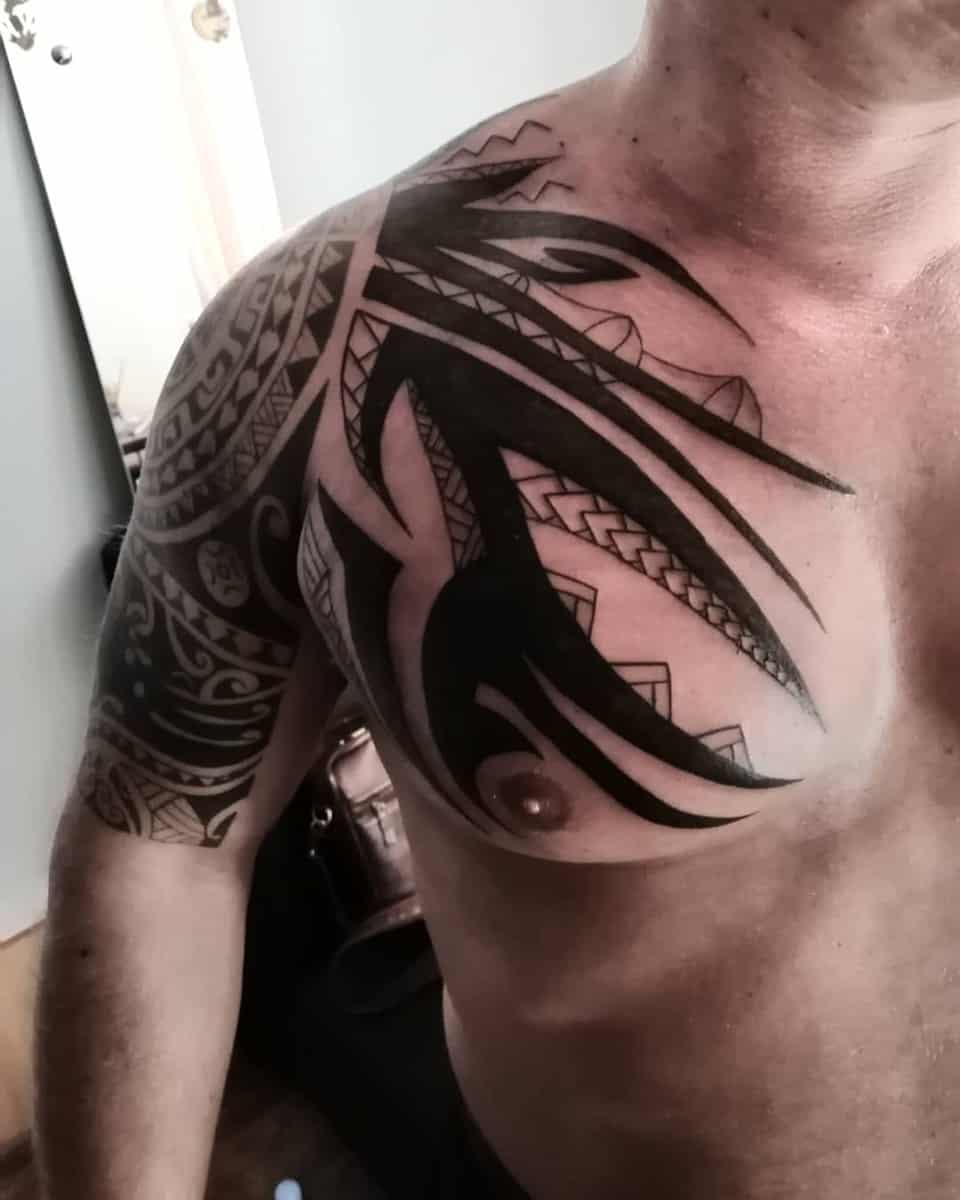 Close-up of a shirtless individual with a large, intricate Polynesian tribal tattoo covering their shoulder, upper arm, and chest