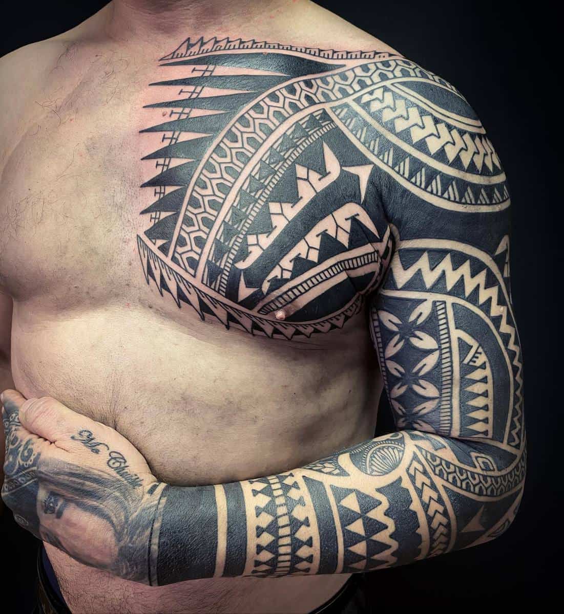 A black Polynesian tattoo covering the right arm, shoulder, and part of the chest, featuring intricate tribal patterns