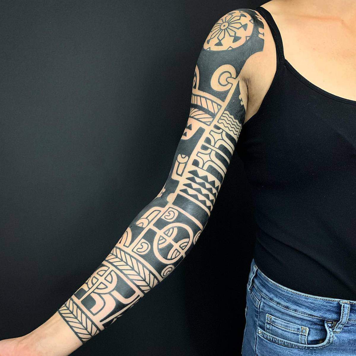 Full sleeve Polynesian tribal tattoo on left arm, wearing a black tank top and blue jeans, against a black background