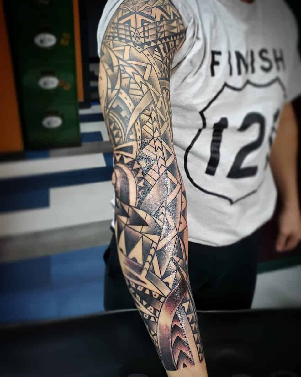 A man wearing a white shirt with 'Finish 12' text displays an intricately designed Polynesian geometric black ink sleeve tattoo on his arm