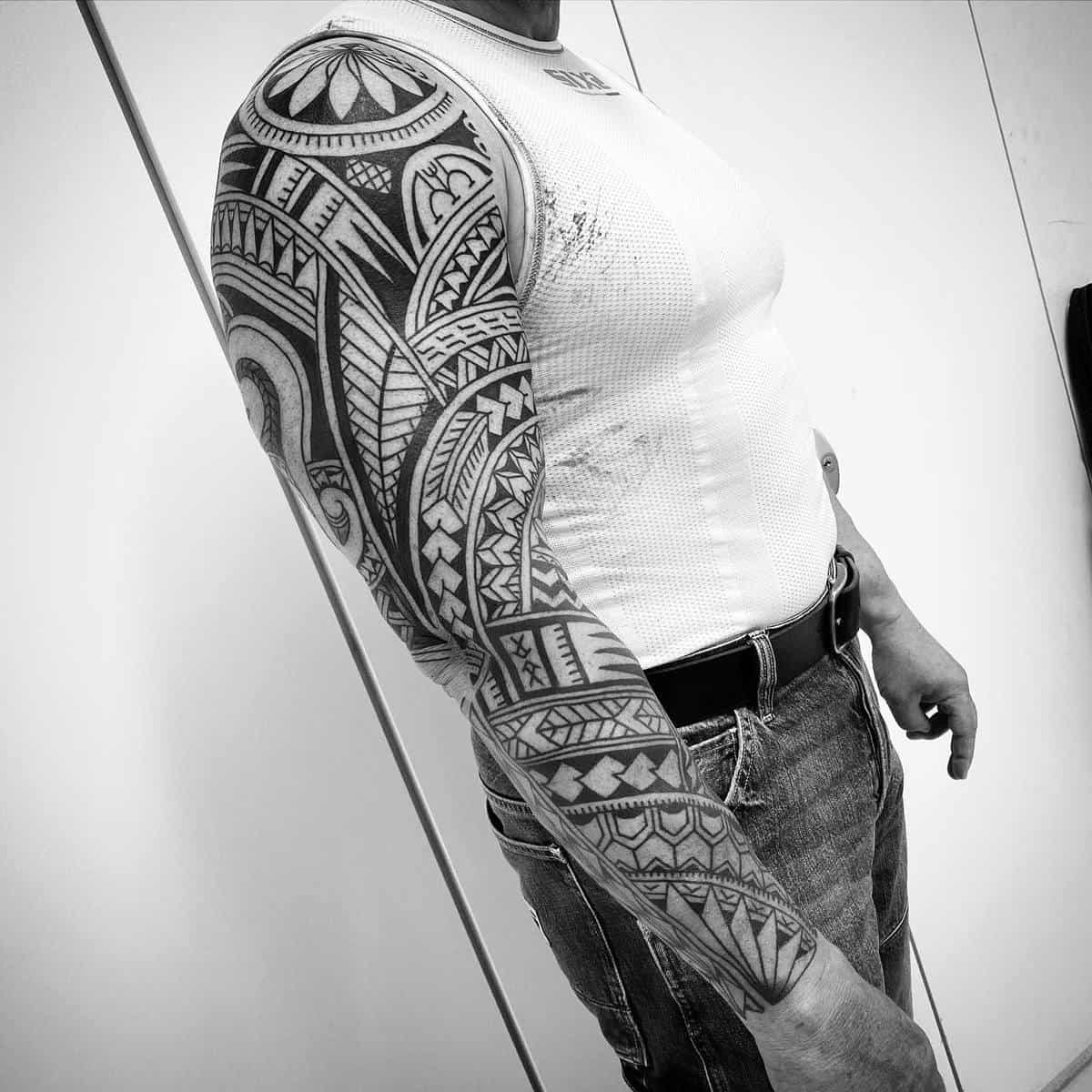 A man in a white sleeveless shirt showcases a detailed Polynesian tribal full-arm tattoo