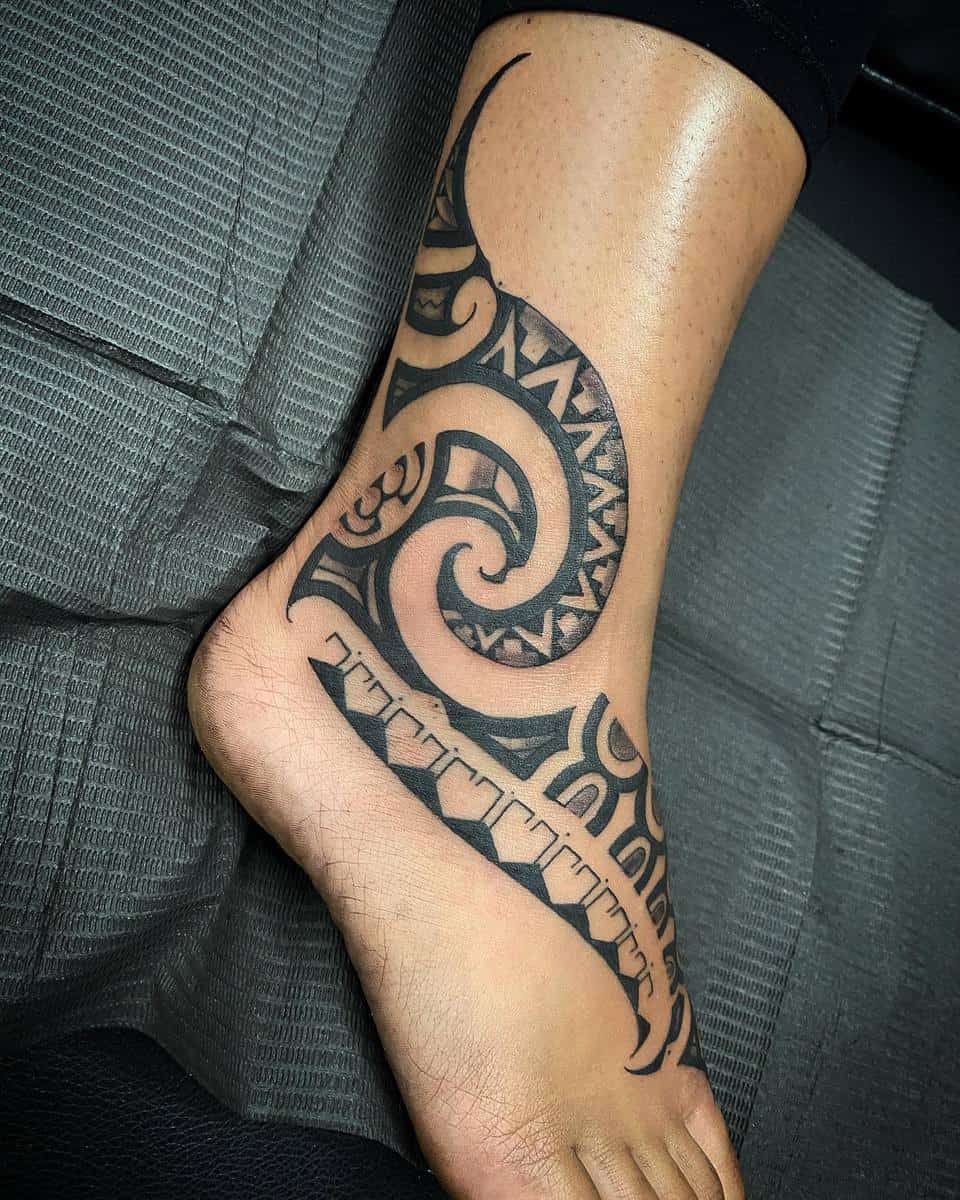 Tattooed foot with intricate tribal design covering the ankle and extending to the toes, seen on a black fabric surface