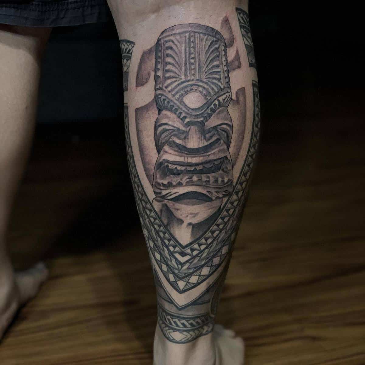A tattoo featuring a detailed tribal mask design on a calf, displayed indoors on a wooden floor