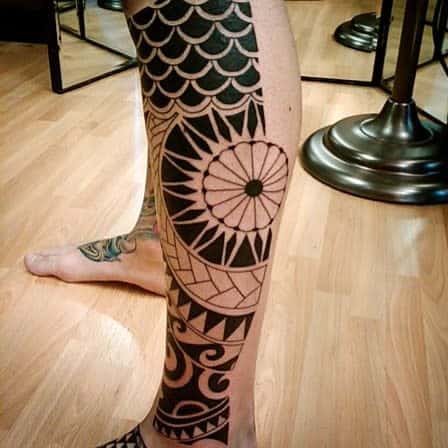 Leg with a large, intricate black geometric tattoo including scales, patterns, and a central circular design against a wood floor
