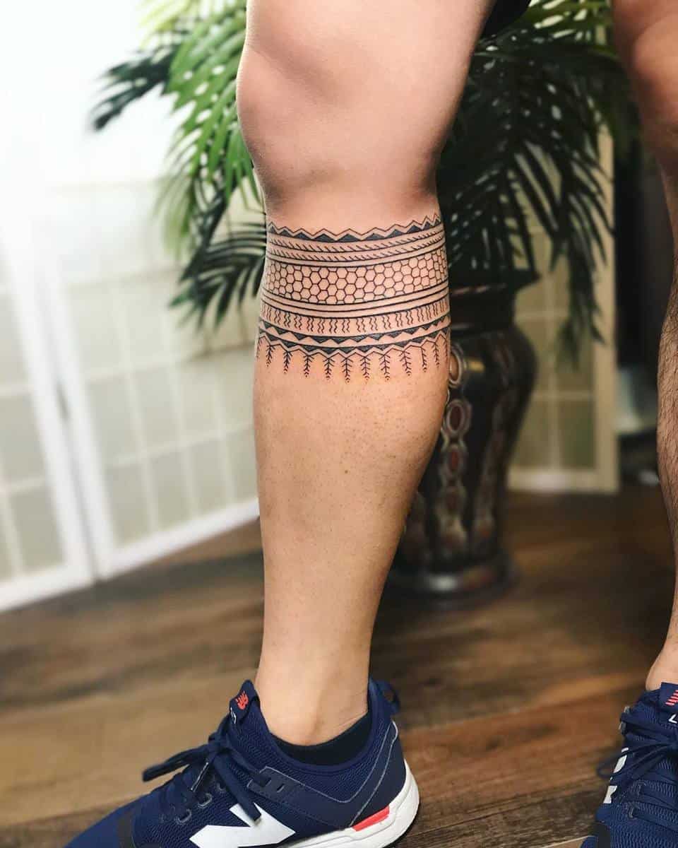 A leg with intricate tribal-style tattoos around the calf, wearing blue sneakers, standing on a wooden floor
