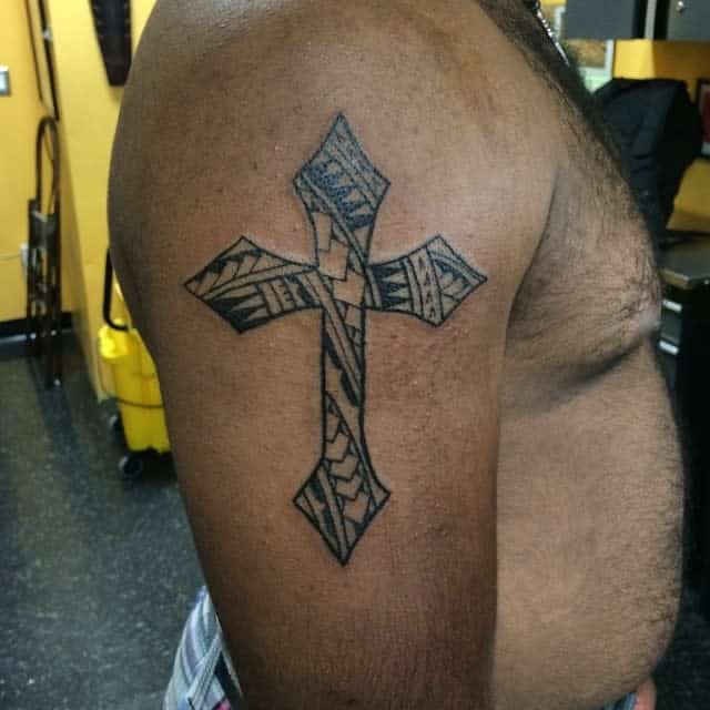 A detailed cross tattoo on an upper arm, standing in a room with yellow walls