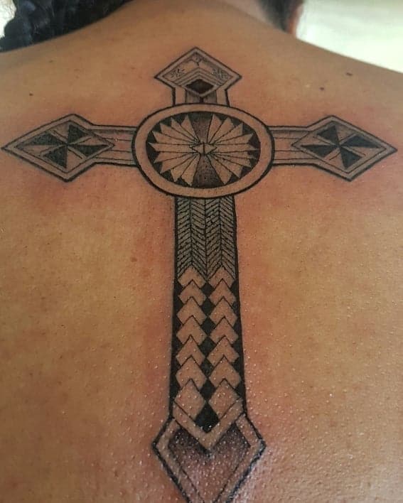 A geometric cross tattoo adorns a person's back, featuring intricate Polynesian patterns and a striking circular design at its center