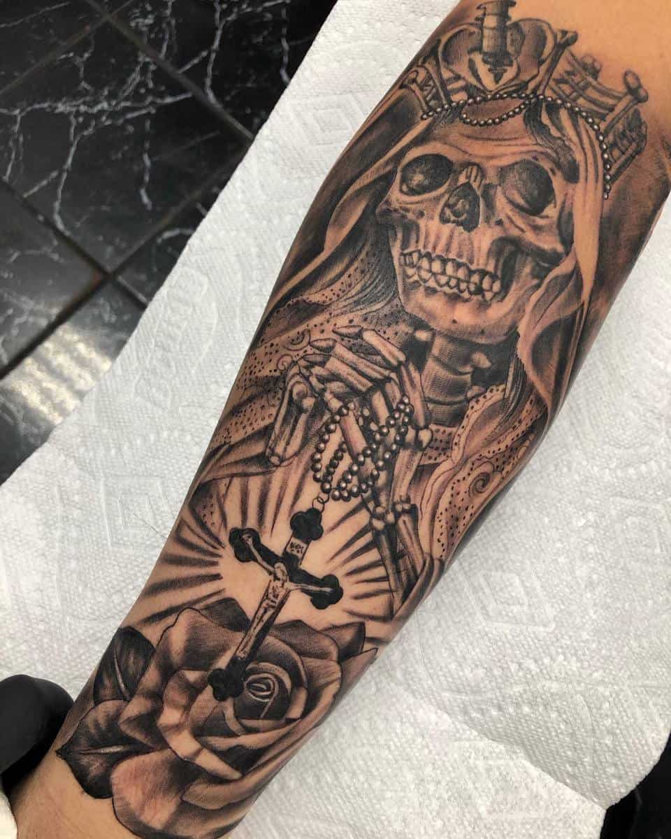 Black-and-grey tattoo of Santa Muerte praying with a rosary, detailed with a crown and a radiant cross atop a rose