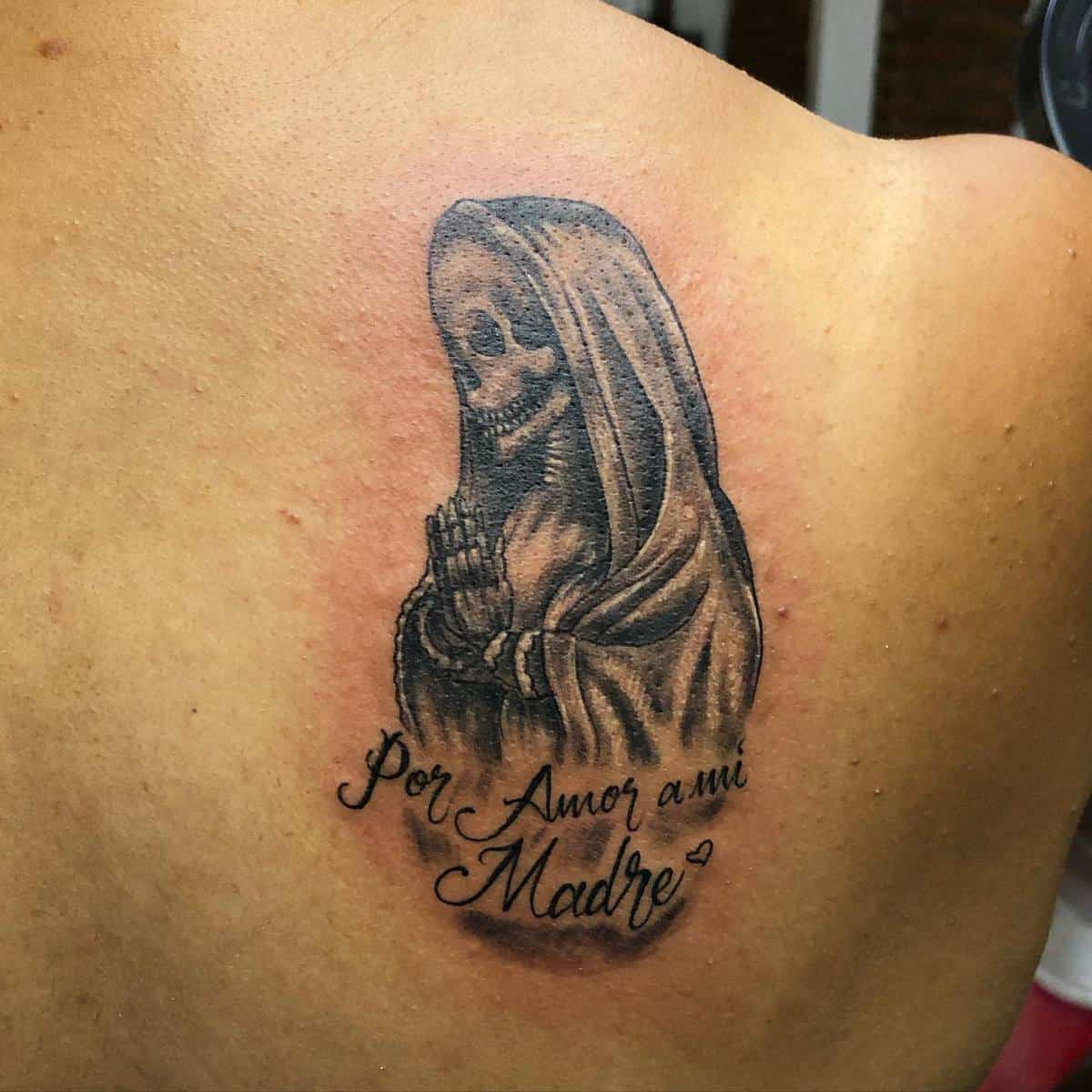A small Santa Muerte tattoo in prayer, with the text "Por Amor a mi Madre" below, symbolizing love for a mother