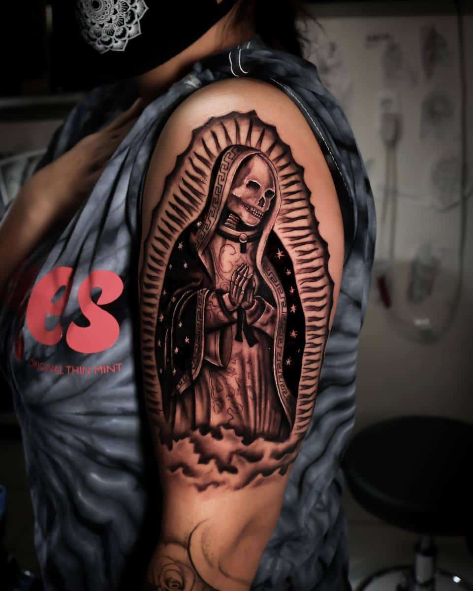 A detailed upper-arm tattoo of Santa Muerte in prayer, wearing a starry robe and framed by radiant lines