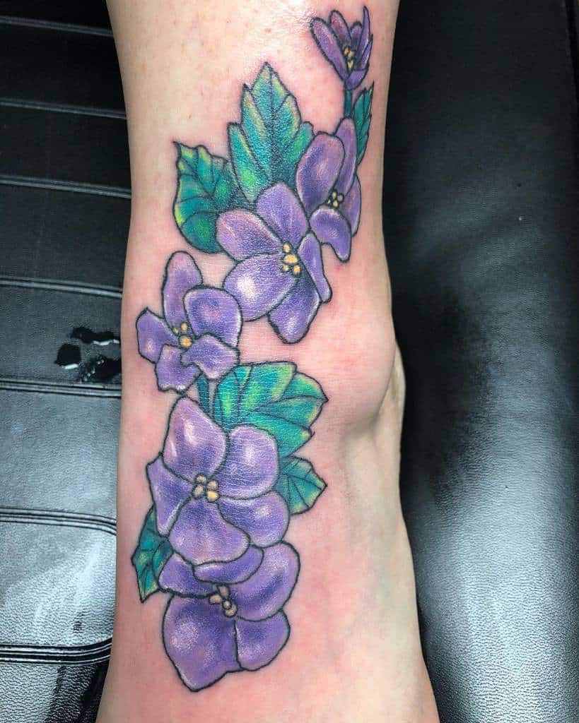 A stunning tattoo of vibrant purple flowers entwined with lush green leaves graces an ankle