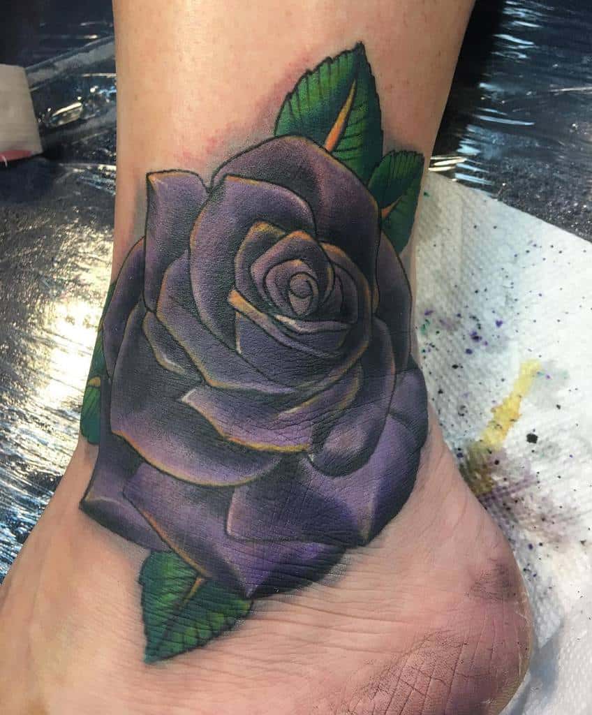 Vibrant purple rose ankle tattoo with green leaves, detailed shading, and a bold design wrapping around the ankle