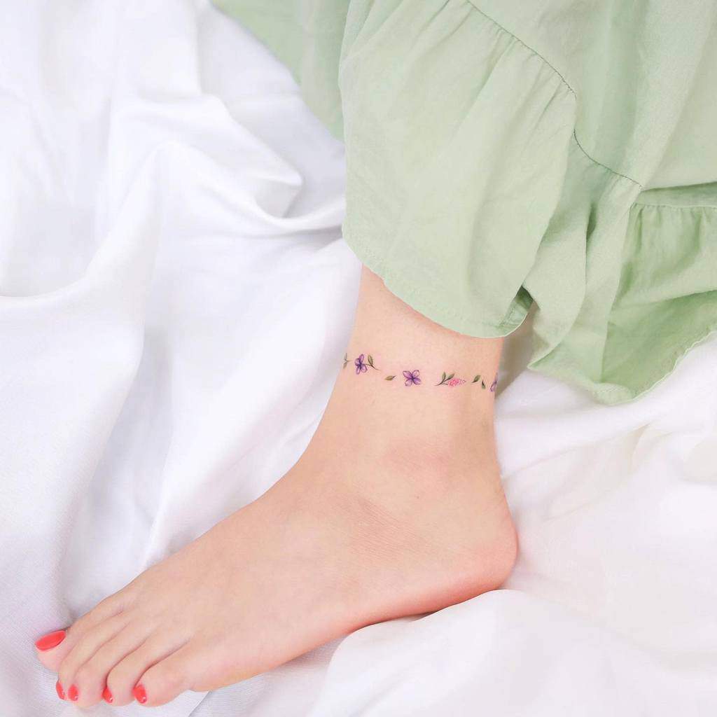 A delicate ankle tattoo of purple flowers and green leaves