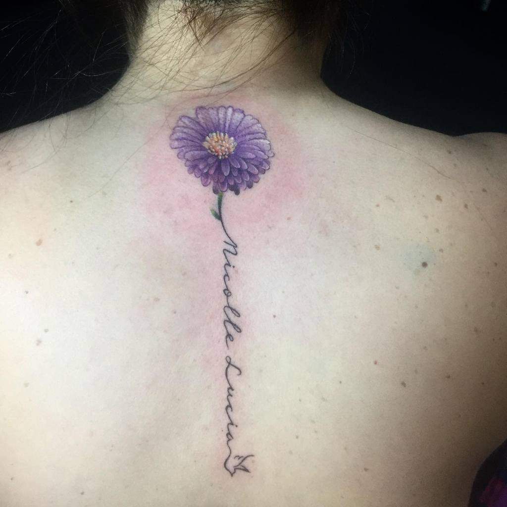 Tattoo of a delicate purple daisy intertwined with elegant script text on an upper back