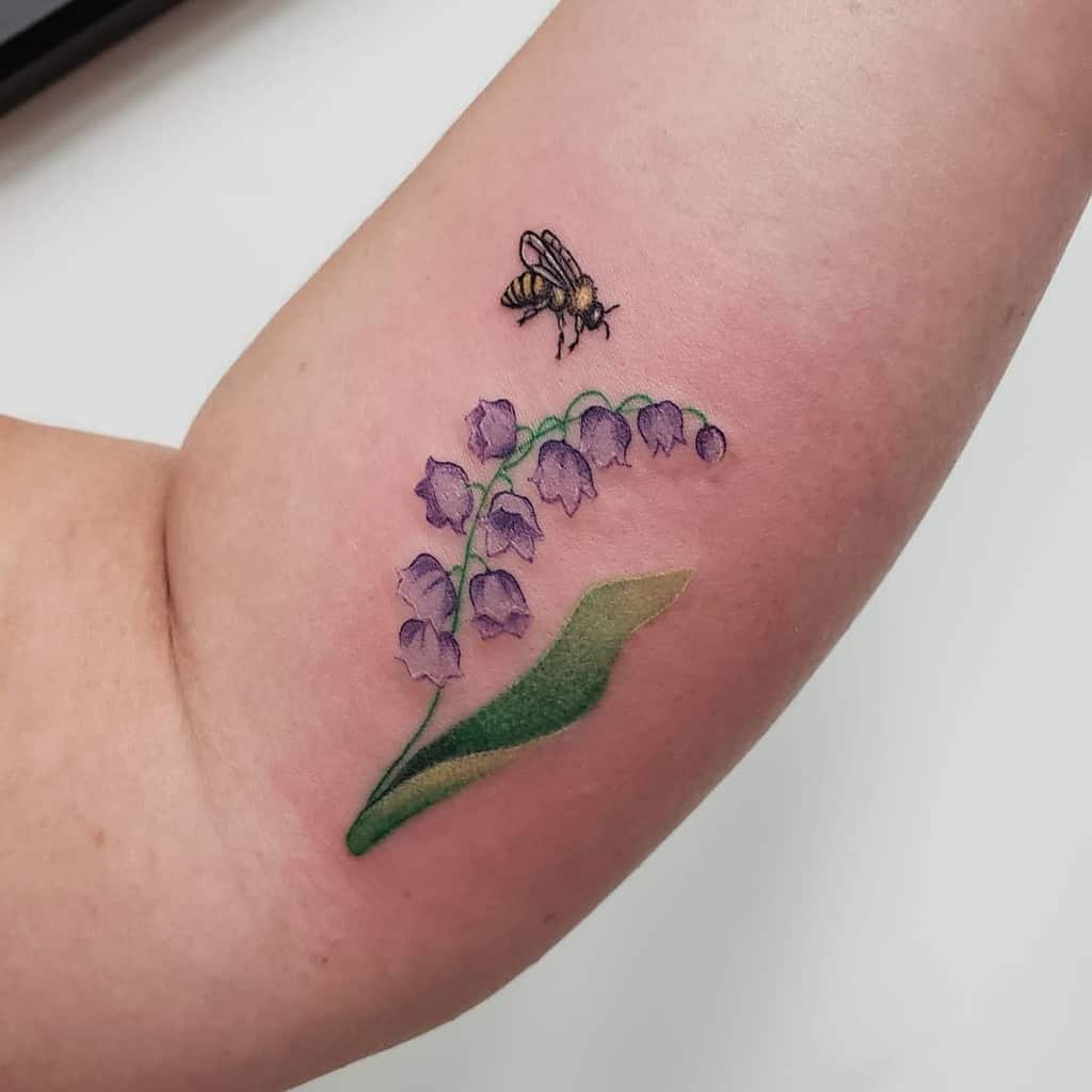 A tattoo of a bee gracefully hovers above vibrant purple lily of the valley flowers, complemented by a lush green leaf on the inner arm