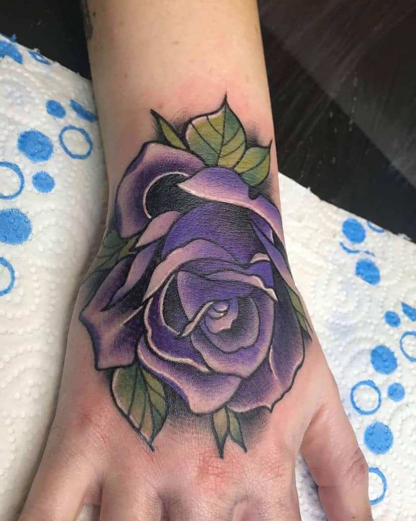 A stunning tattoo of a vibrant purple rose with lush green leaves on a hand