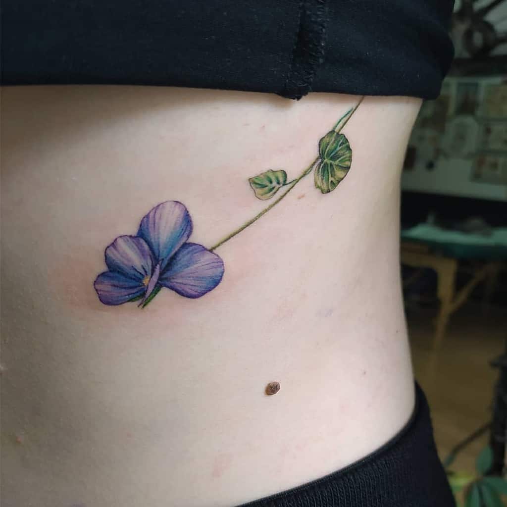 A delicate floral tattoo featuring a vibrant purple flower with lush green leaves graces on the side waist
