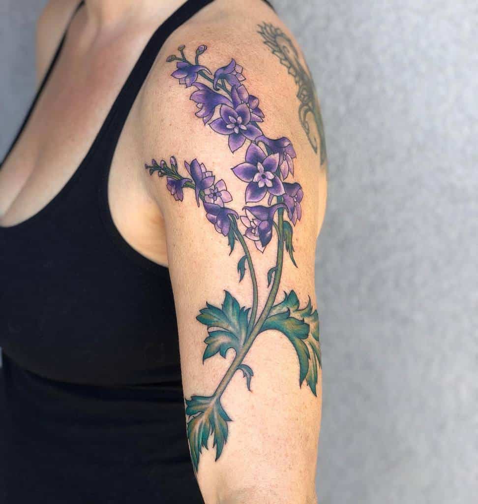 Delicate purple flower tattoo extending from the shoulder down the arm, with vibrant green leaves and detailed shading