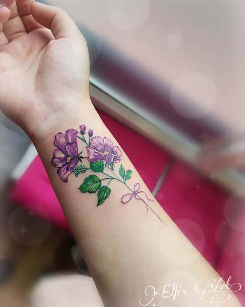 Delicate purple flower wrist tattoo with green leaves and a pink ribbon detail, featuring soft shading and vibrant colors