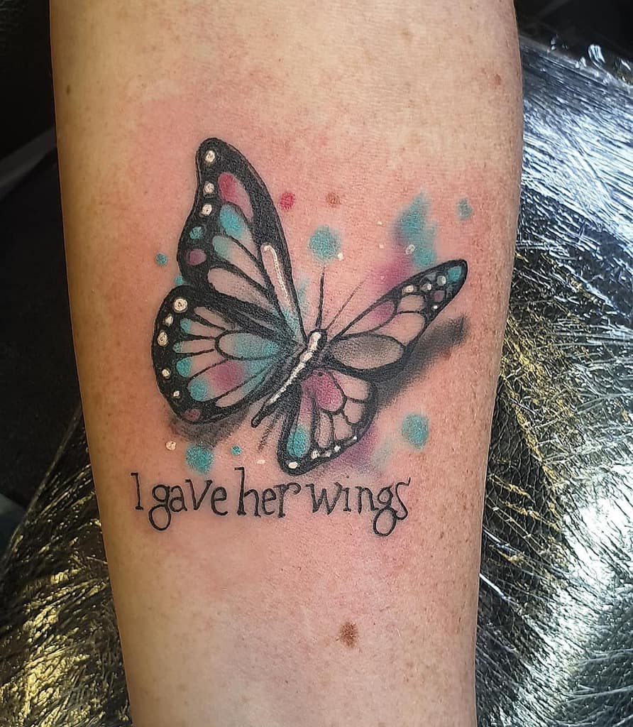 Tattoo of a colorful butterfly with text "I gave her wings" on a person's forearm