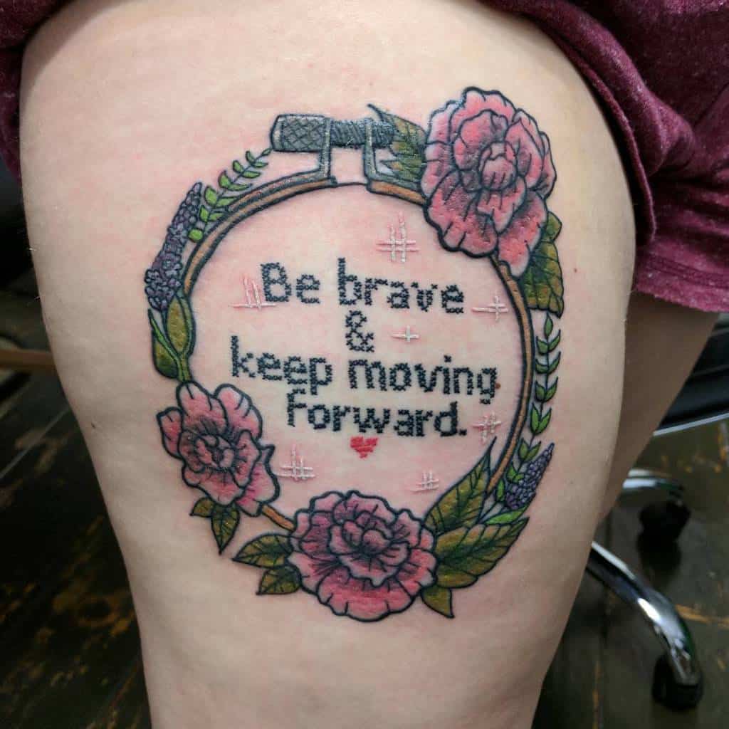 Cross-stitch tattoo of an embroidery hoop with 'Be brave & keep moving forward' text, surrounded by pink roses and leaves for a handmade look