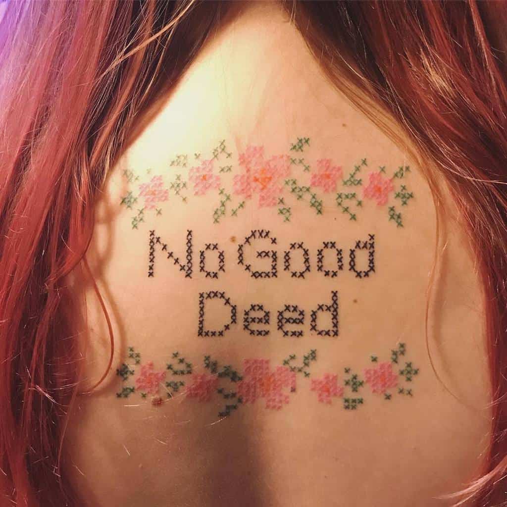 Cross-stitch tattoo featuring 'No Good Deed' in pixelated embroidery-style font, framed by delicate pink floral patterns