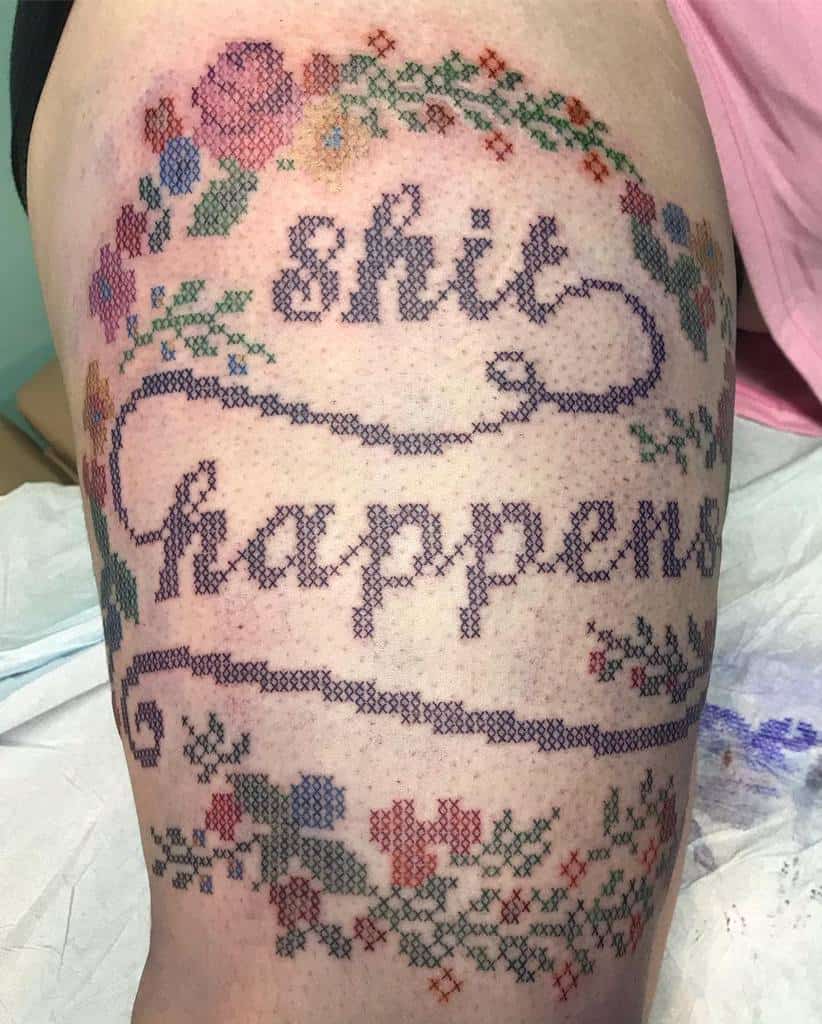 Cross-stitch tattoo with 'Shit Happens' in embroidered-style lettering, surrounded by colorful floral patterns for a vintage, handcrafted look