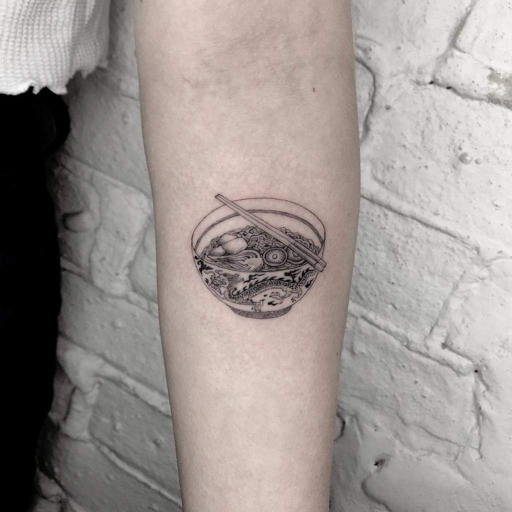 A forearm tattoo of a bowl of ramen with chopsticks, intricately designed using single needle technique, featuring various ingredients