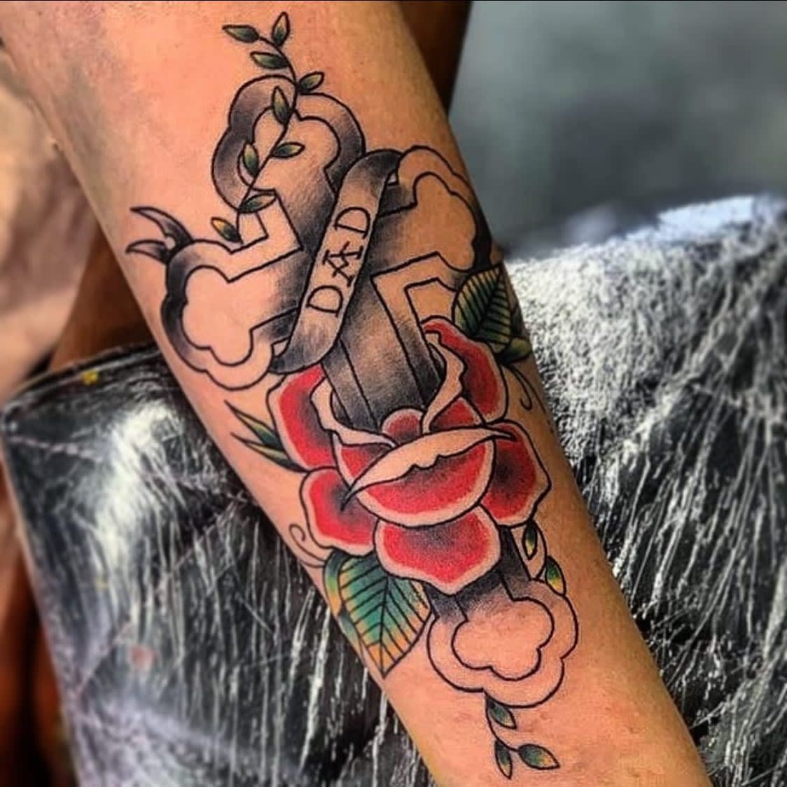 Tattoo of a cross with "Dad" on it, surrounded by flowers and leaves, on a forearm