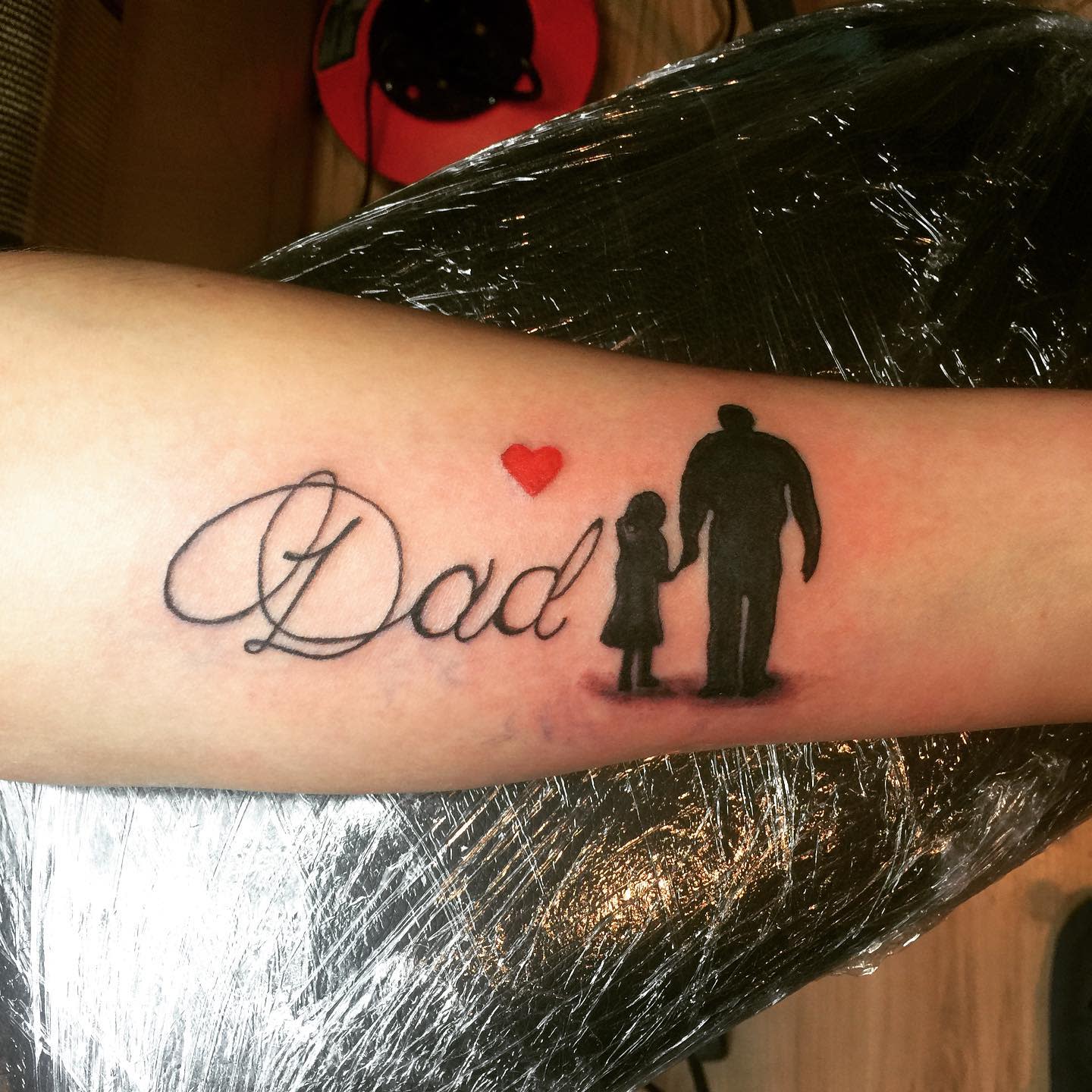 Tattoo of "Dad" with a heart and silhouette of a man holding a child's hand on forearm