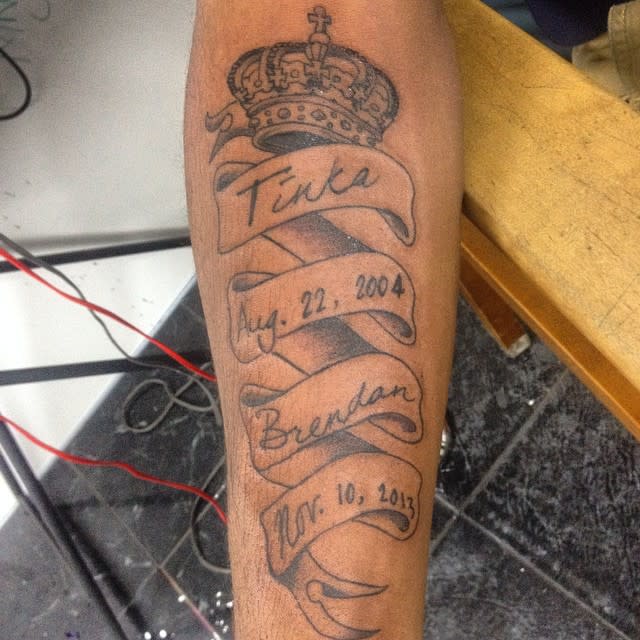 Forearm tattoo of a crown with names and dates written on a ribbon below