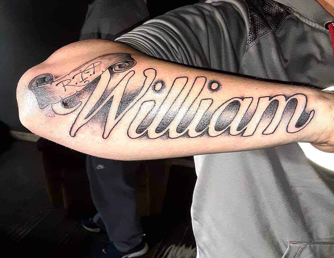 Forearm tattoo with the name "William" and a small banner above it reading "R.I.P."