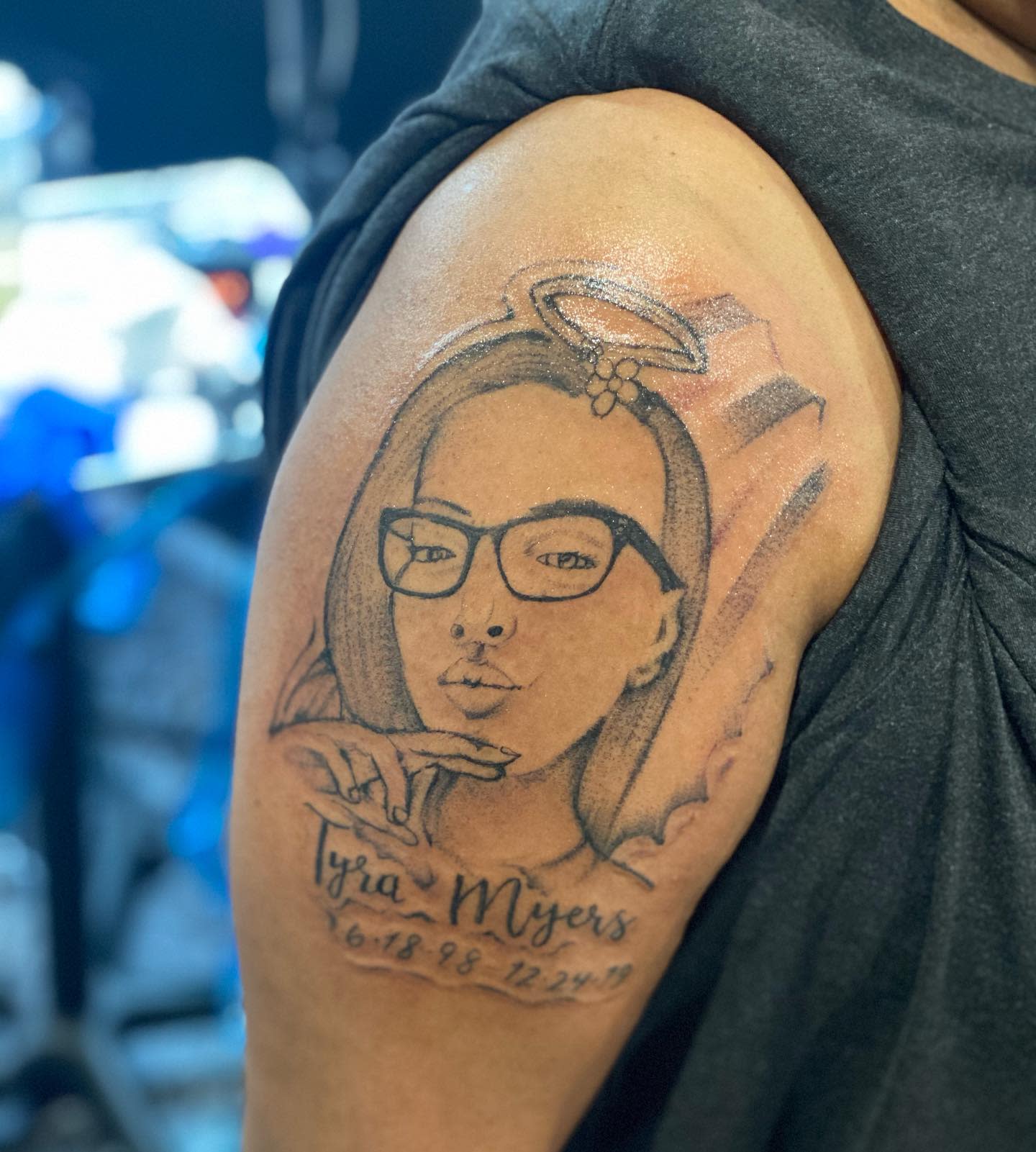 Memorial tattoo of a woman with glasses and halo, dates, and "Tyra Myers" on the upper arm