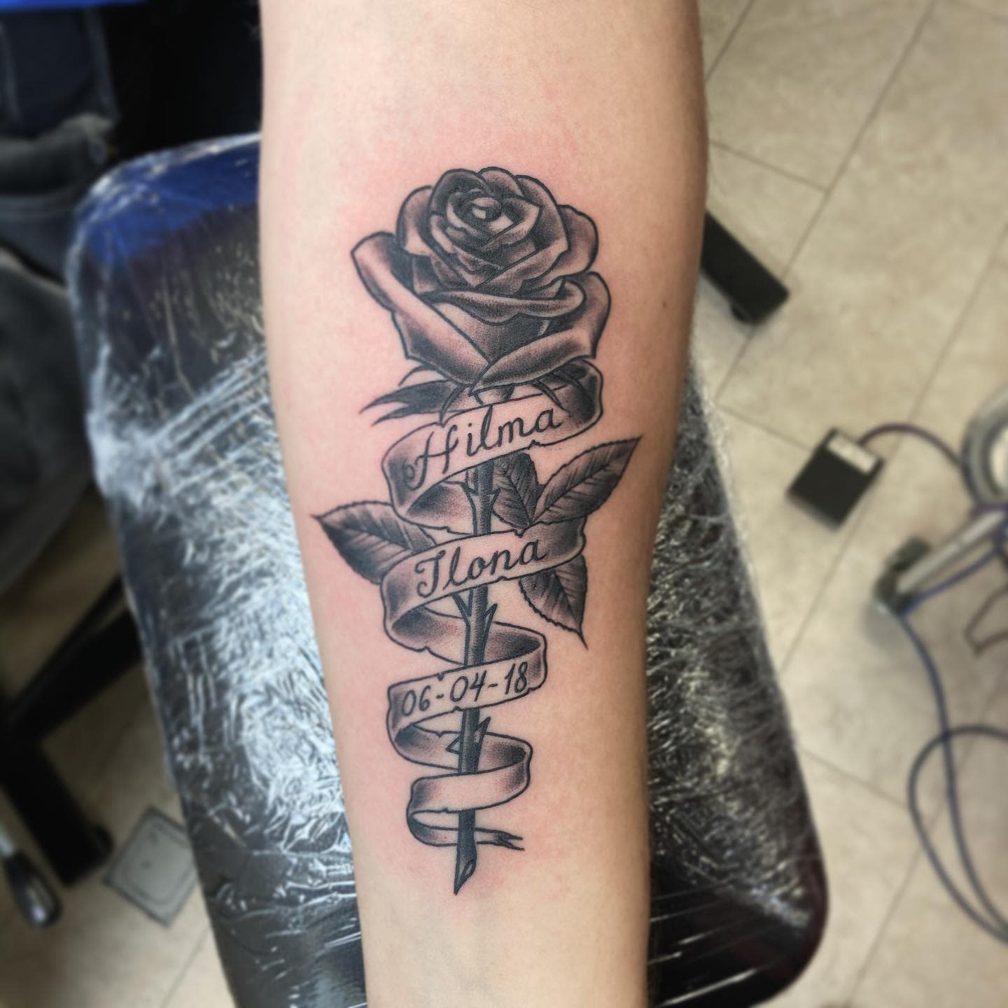Tattoo of a rose with leaves with names "Hilma" and "Ilona" and date "06-04-18" on forearm