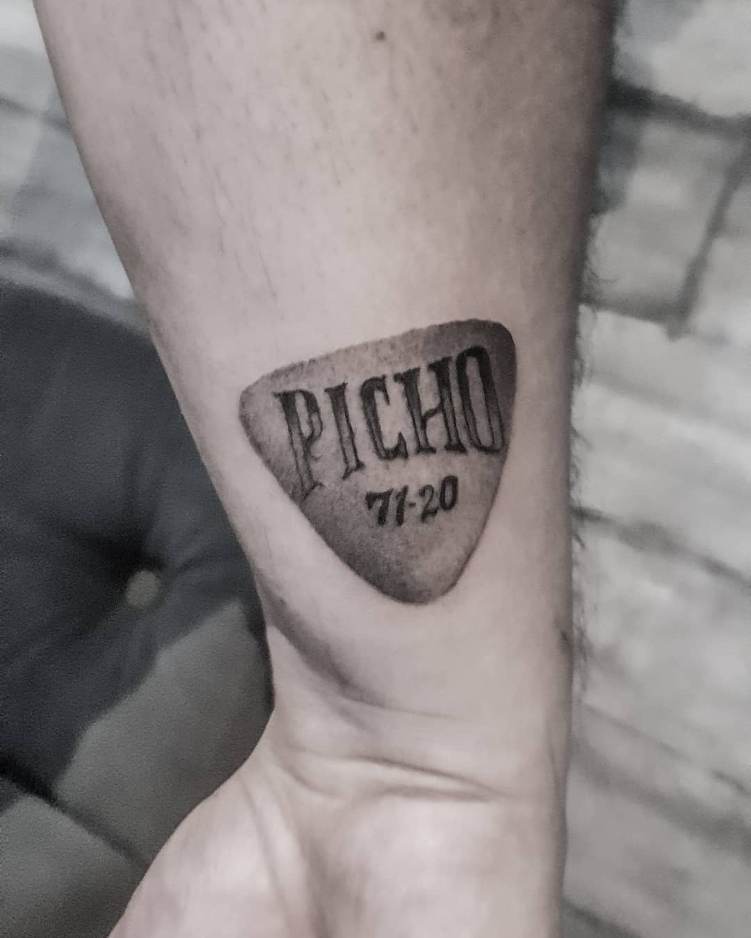 Tattoo of a guitar pick with "PICHO 71-20" on a wrist