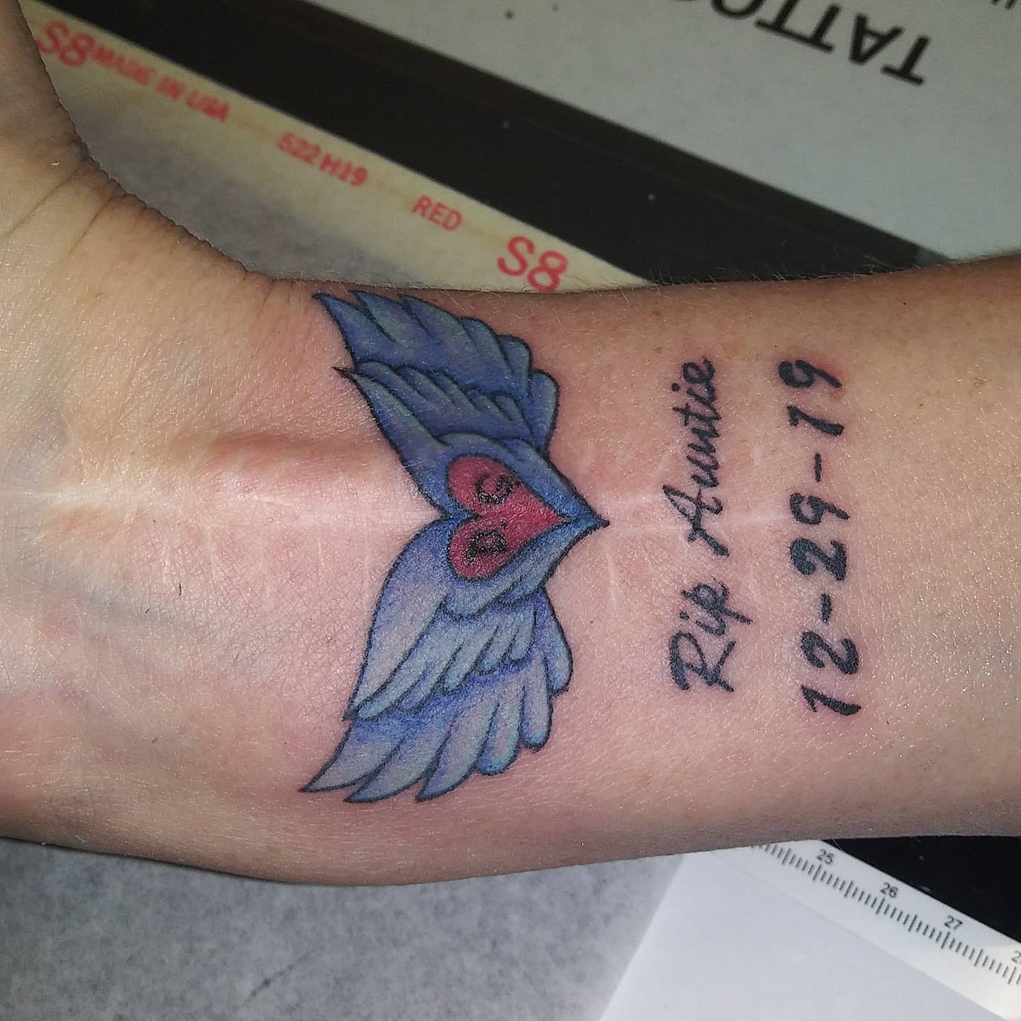 Tattoo of a heart with wings on a wrist, text reads "Rip Auntie 12-29-19"