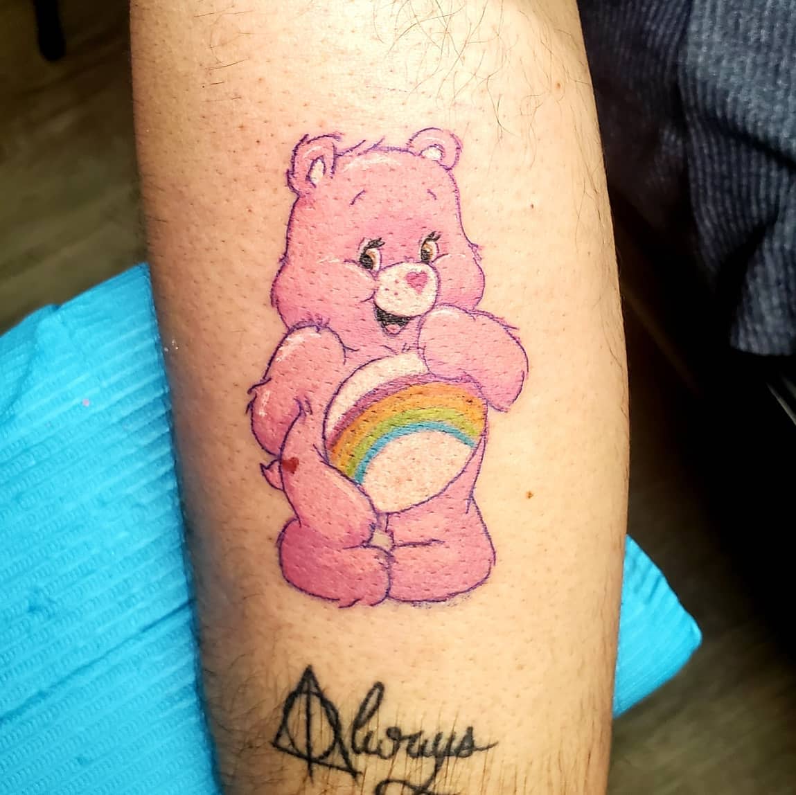 Tattoo of a pink bear holding a rainbow on an arm with a signature below