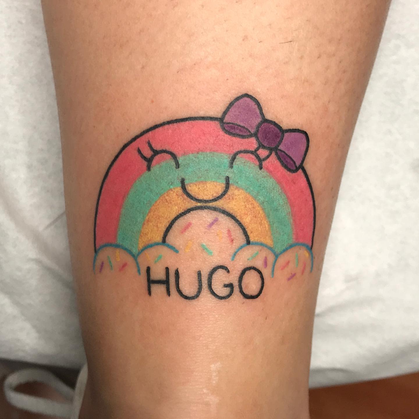 Tattoo of a smiling rainbow with a bow and the name "Hugo" written below