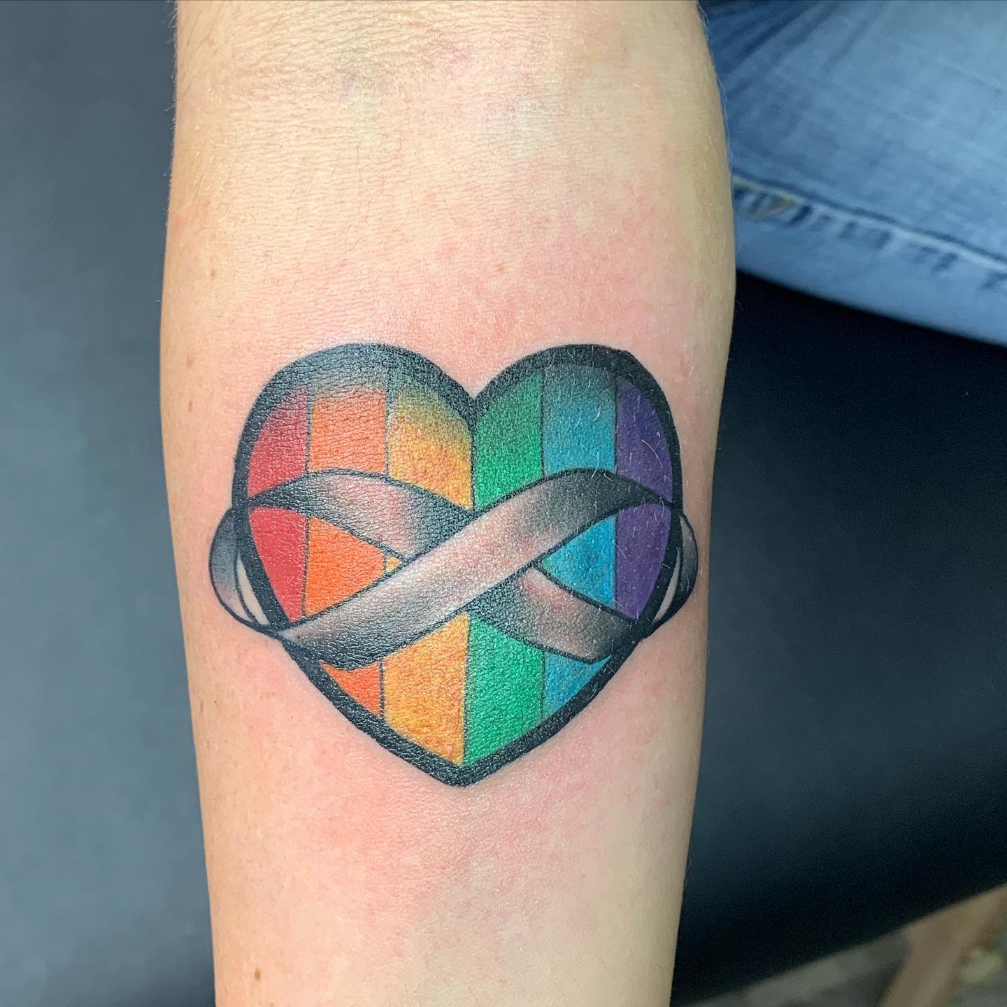 Tattoo of a heart with rainbow stripes and an infinity symbol on an arm