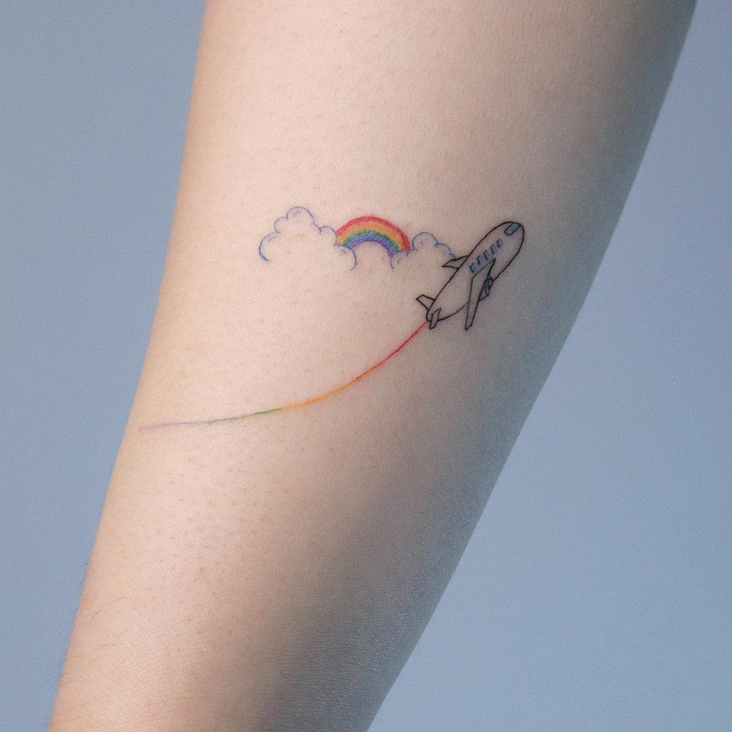 Tattoo of a plane flying through clouds with a rainbow trailing behind on an arm
